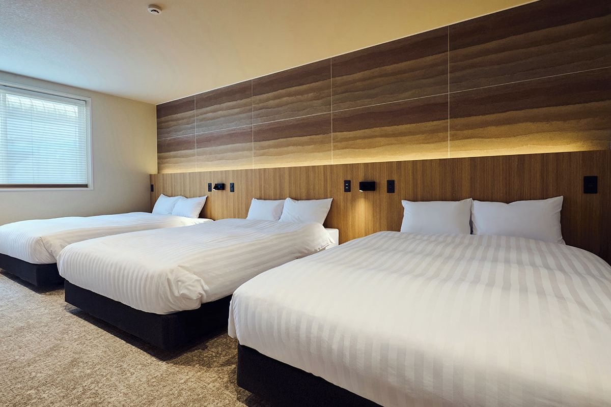 A hotel room with three beds, a large window, and a modern wooden headboard.
