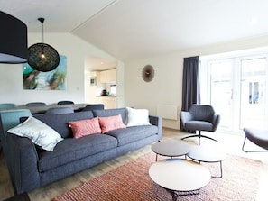 Ein modernes Wohnzimmer mit einem grauen Sofa, einem Esstisch und einem Stuhl. Durch einen offenen Durchgang ist eine Küche erkennbar.