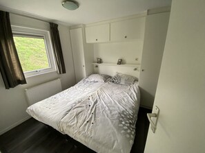 Ein Schlafzimmer mit einem Einzelbett, einem fensterbelaubten Fenster, einem Heizkörper und einer weißen Wand mit einem Regal über dem Bett.