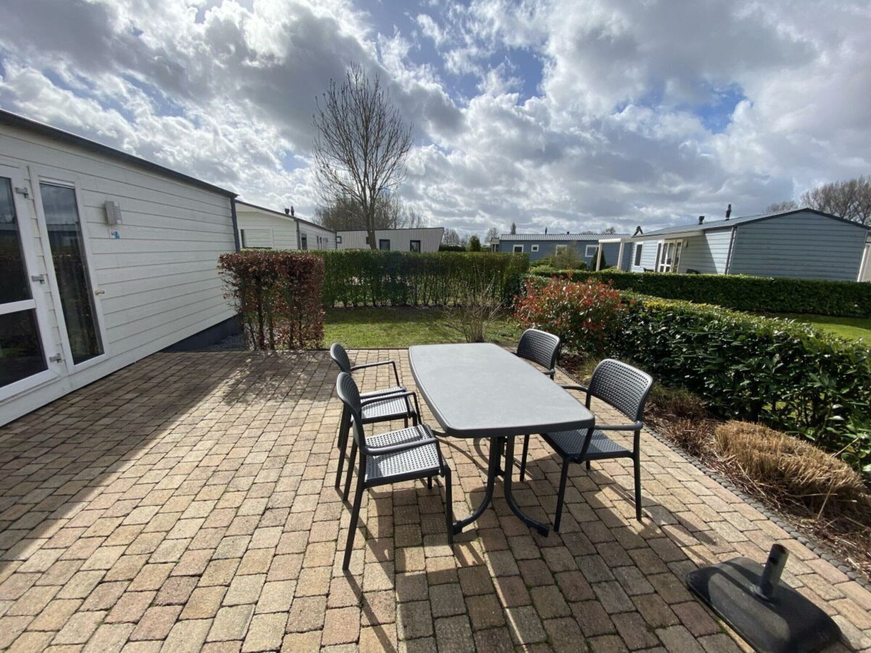 A patio with a table and chairs, a hedge, and a row of mobile homes in the background.