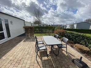 Eine Terrasse mit Tisch und Stühlen, ein Hecken und eine Reihe von Mobilheimen im Hintergrund.