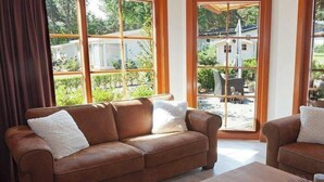 Un canapé en cuir marron avec des coussins blancs, une table basse en bois et de grandes fenêtres donnant sur un jardin et une maison.