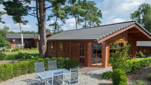 Une cabane en bois avec un toit en tuiles, une terrasse avec une table et des chaises, et un jardin agrémenté de haies et d’arbres.