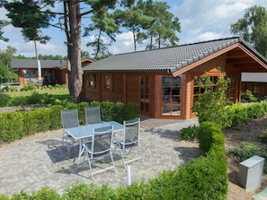Eine Holzhütte mit Ziegeldach, einer Terrasse mit Tisch und Stühlen sowie ein Garten mit Hecken und Bäumen.