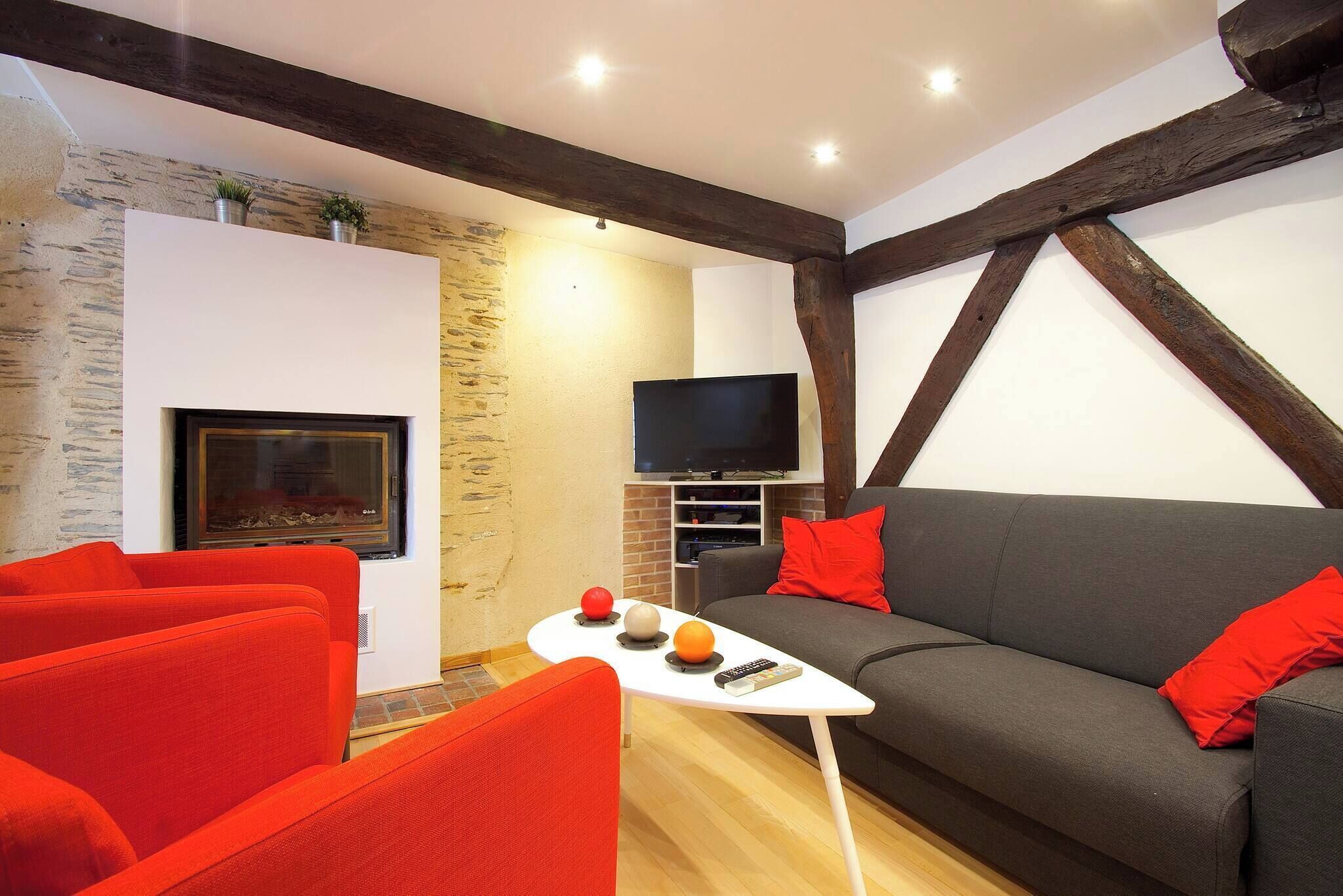 A living room with a fireplace, a television, a sofa with red cushions, and a small table with fruits and a remote control.