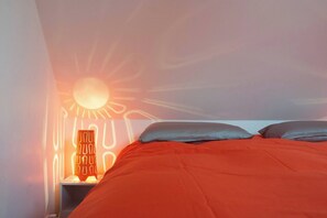 A bed with a red blanket, a nightstand with a lamp, and a wall projection of a sun. - AppartCosy 02  Rachapt Duplex 30m2 (3 Pers) (Vitré)