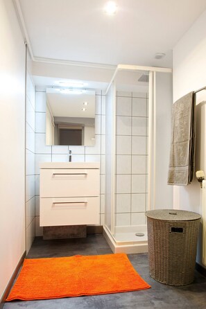 A bathroom with a white vanity, a shower enclosure, and a wicker basket. - AppartCosy 02  Rachapt Duplex 30m2 (3 Pers) (Vitré)