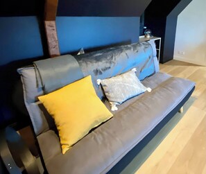 A grey sofa with two cushions, one yellow and one grey, in a room with a blue accent wall and a wooden floor. - AppartCosy 15 ArtDéco (Vitré)