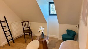 Una habitación con techo inclinado, una silla de madera, una mesita con un florero y una silla color turquesa, además de una escalera apoyada en un radiador.