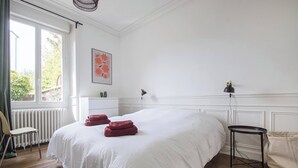 A bedroom with a large bed, a chair, a small table, a radiator, and a window. - AppartCosy 09 for 4 people in a charming house (Vitré)