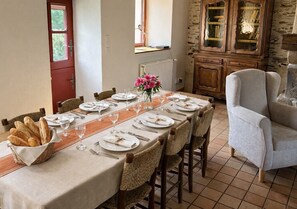 Une salle à manger avec une longue table dressée pour six personnes, entourée de chaises en osier, d’un buffet aux portes vitrées et d’une porte rouge.