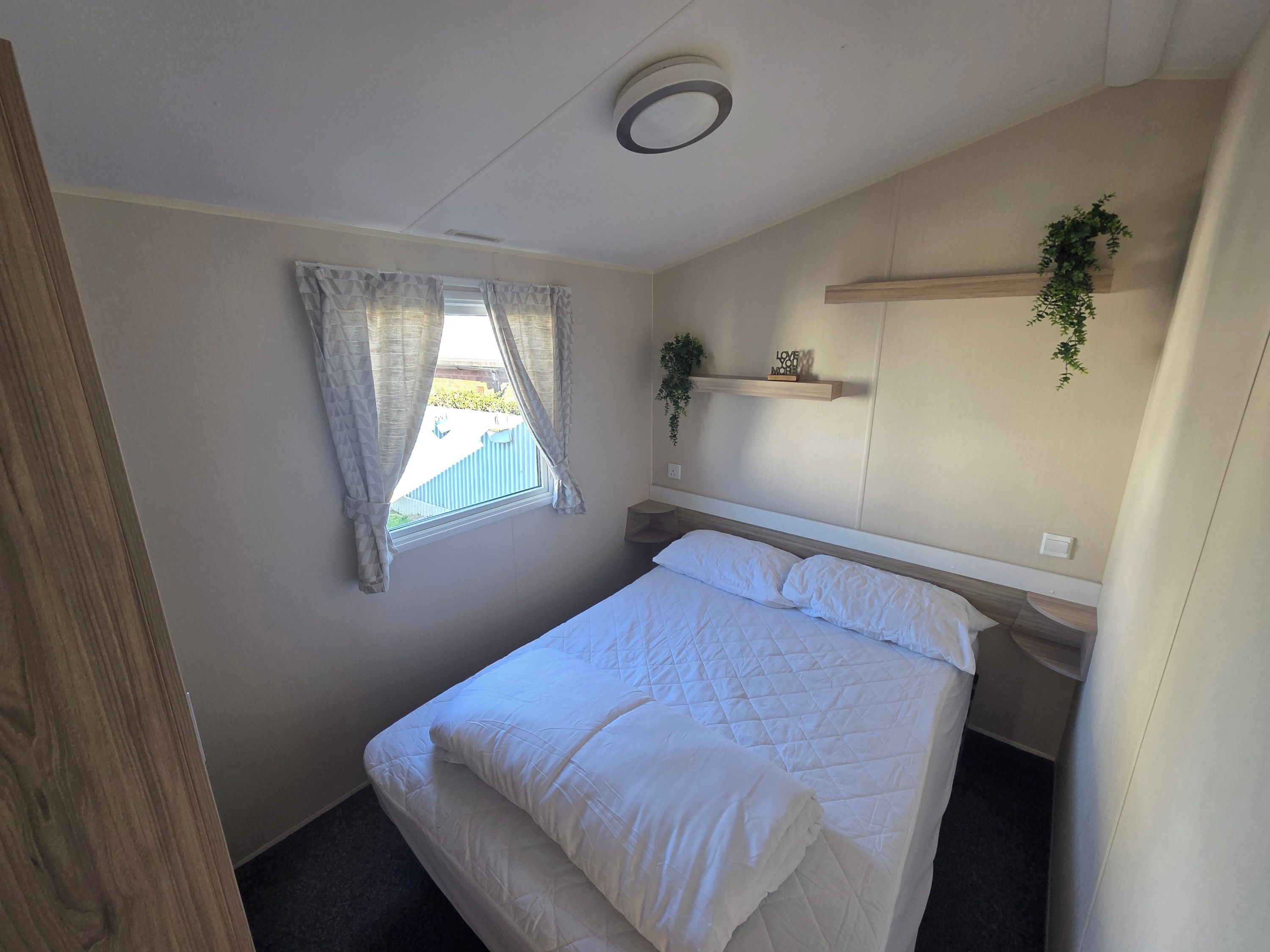 A bedroom with a bed, a window with curtains, a bedside table, a shelf with a plant, and a ceiling light.