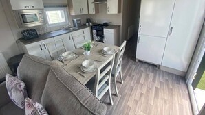 Een compacte keuken met een magnetron, broodrooster en fornuis, een eethoek met een tafel gedekt voor twee personen, en een grote witte kast.