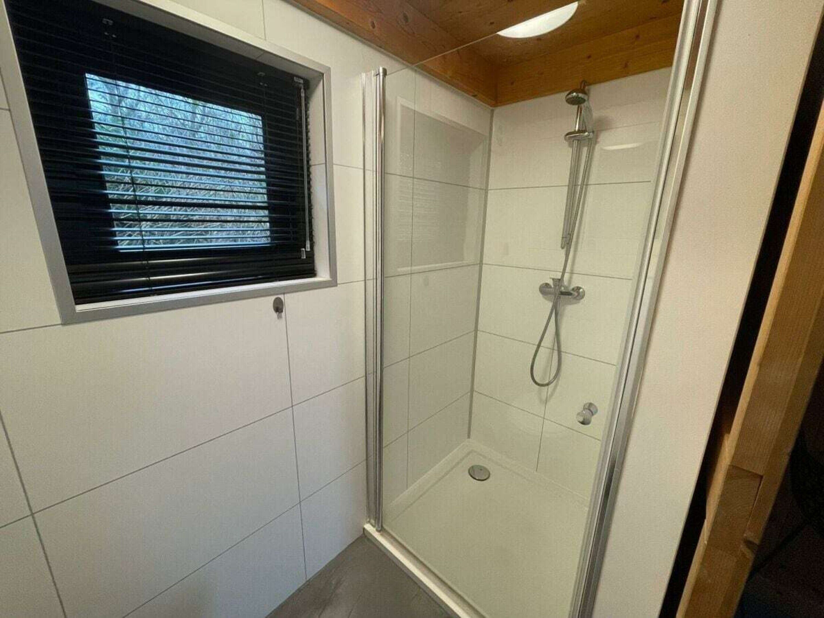 A glass-enclosed shower with a handheld showerhead, a window with blinds, and white tiled walls.