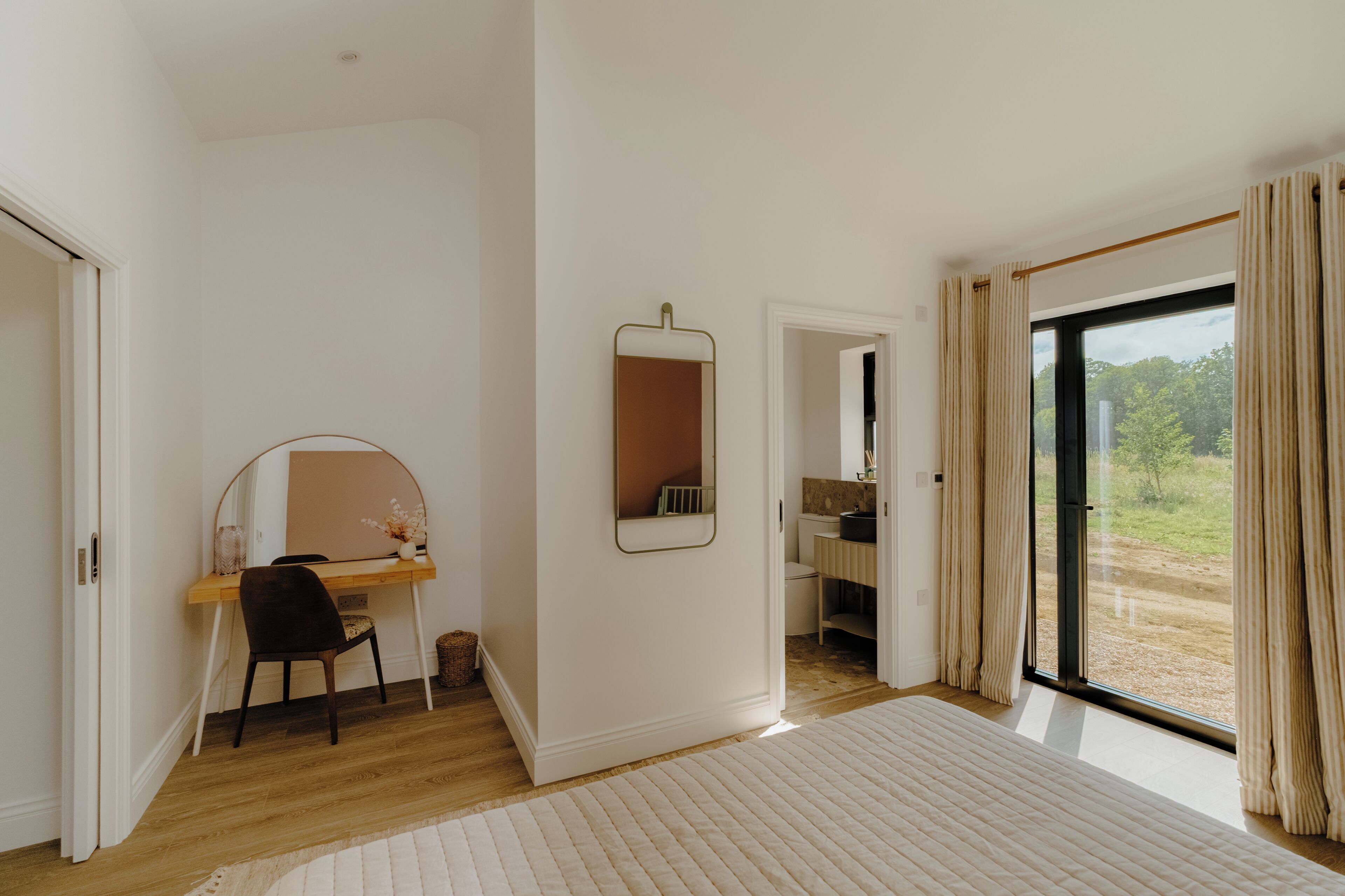 Een slaapkamer met een houten bureau, een spiegel, een bed en uitzicht door een groot raam.
