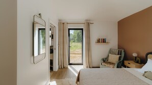 Ein Schlafzimmer mit Bett, Stuhl, Nachttisch, Spiegel und Blick durch eine Tür auf einen Außenbereich.