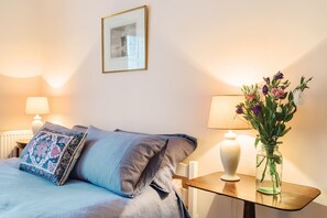 A neatly made bed with a blue bedspread and decorative pillows, a small table with a vase of flowers, and two wall-mounted lamps. - Artist's Studio (2) - Piddlehinton, Dorchester, sleeps 2 guests  in 1 bedroom (Piddlehinton)