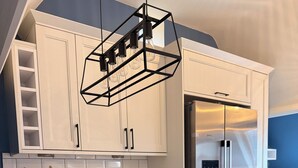 Une cuisine moderne avec des meubles blancs, un réfrigérateur en acier inoxydable, un luminaire au design géométrique original et un coin petit-déjeuner avec des croissants et du jus d’orange.