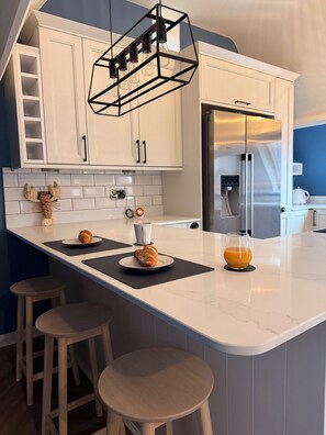 Eine moderne Küche mit weißen Schränken, einem Edelstahlkühlschrank, einem außergewöhnlichen geometrischen Lichtobjekt und einer Frühstücksecke mit Croissants und Orangensaft.