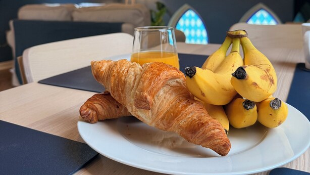 Une table dressée pour le petit-déjeuner avec des croissants et des bananes sur une assiette, un verre de jus d’orange et une vue sur une pièce avec une fenêtre et une lampe.