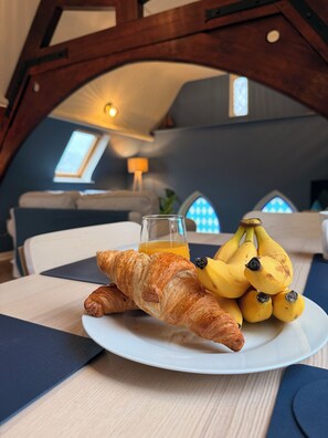 Ein Frühstückstisch mit Croissants und Bananen auf einem Teller, ein Glas Orangensaft und Blick in einen Raum mit Fenster und Lampe.