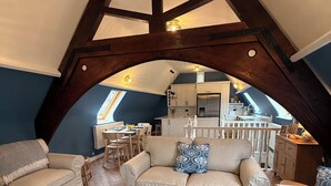 Une chambre située sous les toits, avec un plafond en pente, des poutres apparentes, un coin cuisine et un espace salon comprenant deux canapés et une table basse.