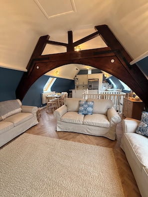 Une chambre située sous les toits, avec un plafond en pente, des poutres apparentes, un coin cuisine et un espace salon comprenant deux canapés et une table basse.