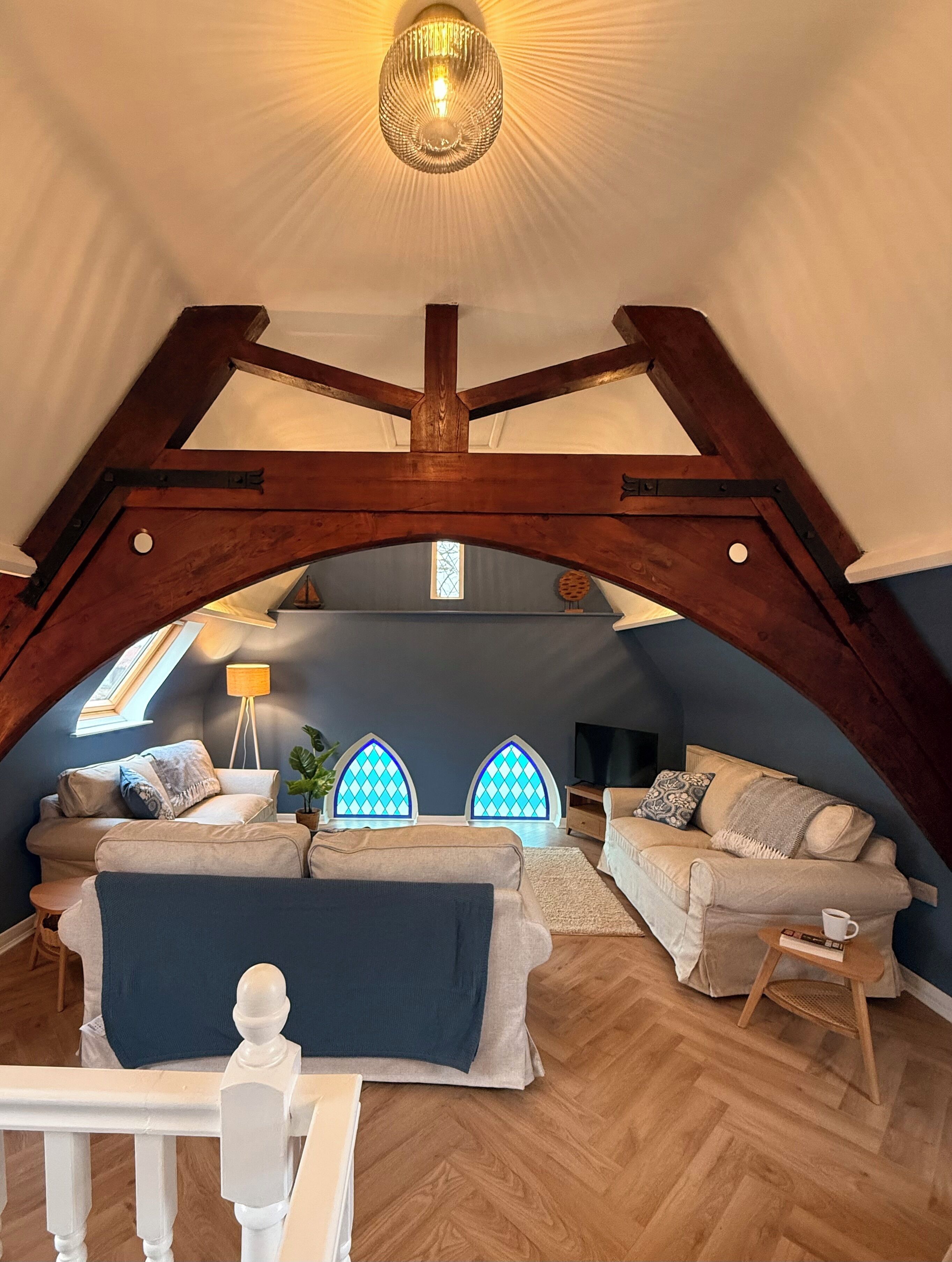 An attic room with exposed wooden beams, a large chandelier, two stained glass windows, and a cozy seating area with sofas and a coffee table.