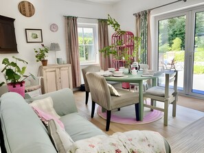 Uma sala de estar aconchegante com sofá, mesa de jantar com cadeiras, uma poltrona rosa e uma janela grande com cortinas.
