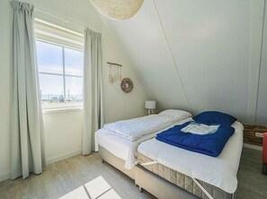 Eine kleine Einzimmerwohnung mit schrägen Wänden, einem Bett mit weißer Bettwäsche und einem blauen Kissen, einem kleinen Tisch mit Lampe, Blick auf die Stadt durch ein großes Fenster und einer Pendelleuchte.