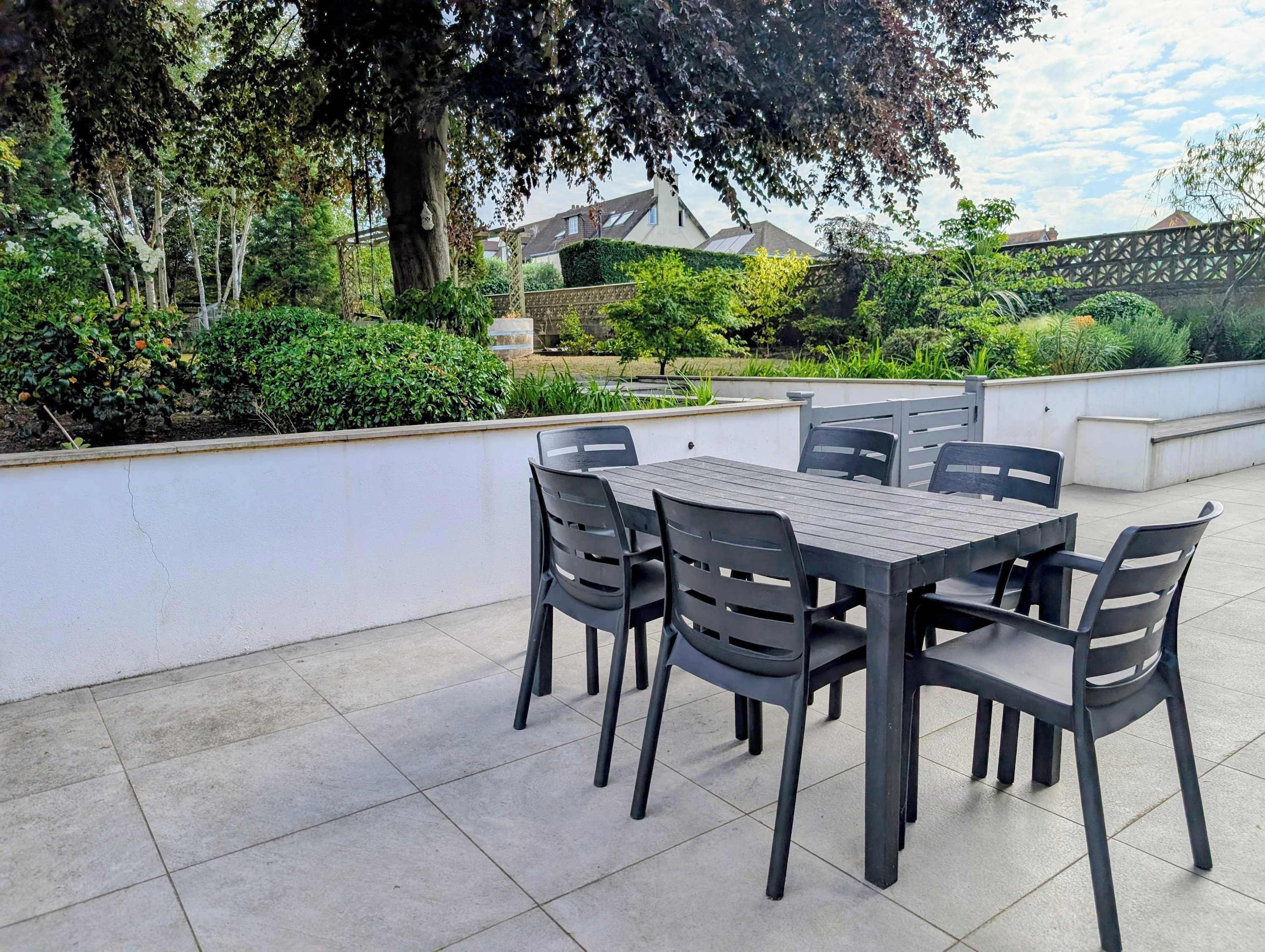 Un patio al aire libre con una mesa y sillas, rodeado de follaje y una pared blanca.