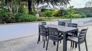 Un patio al aire libre con una mesa y sillas, rodeado de follaje y una pared blanca.