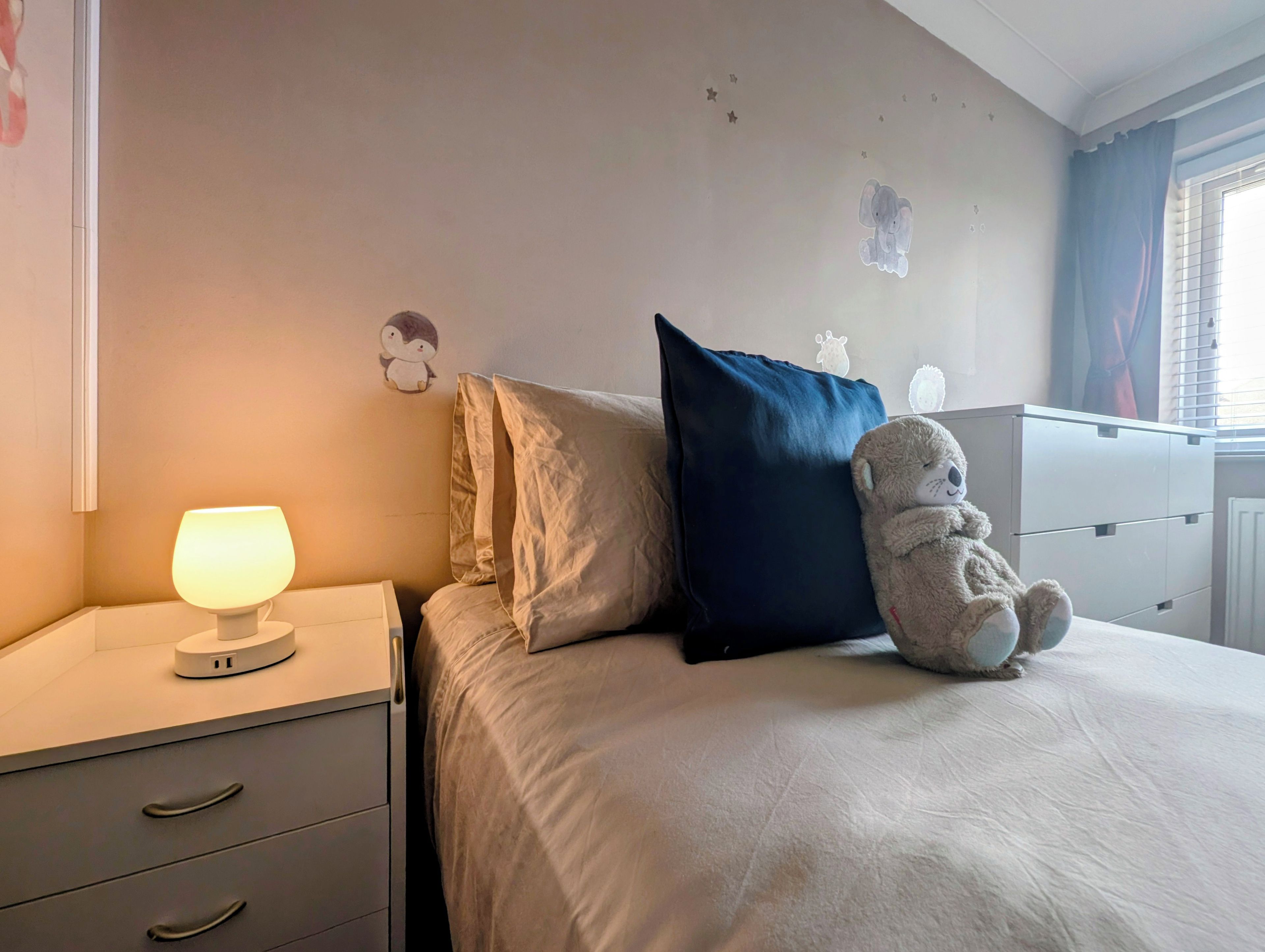 Un dormitorio con una cama, una lámpara de noche, un osito de peluche y un aparador con cajones.