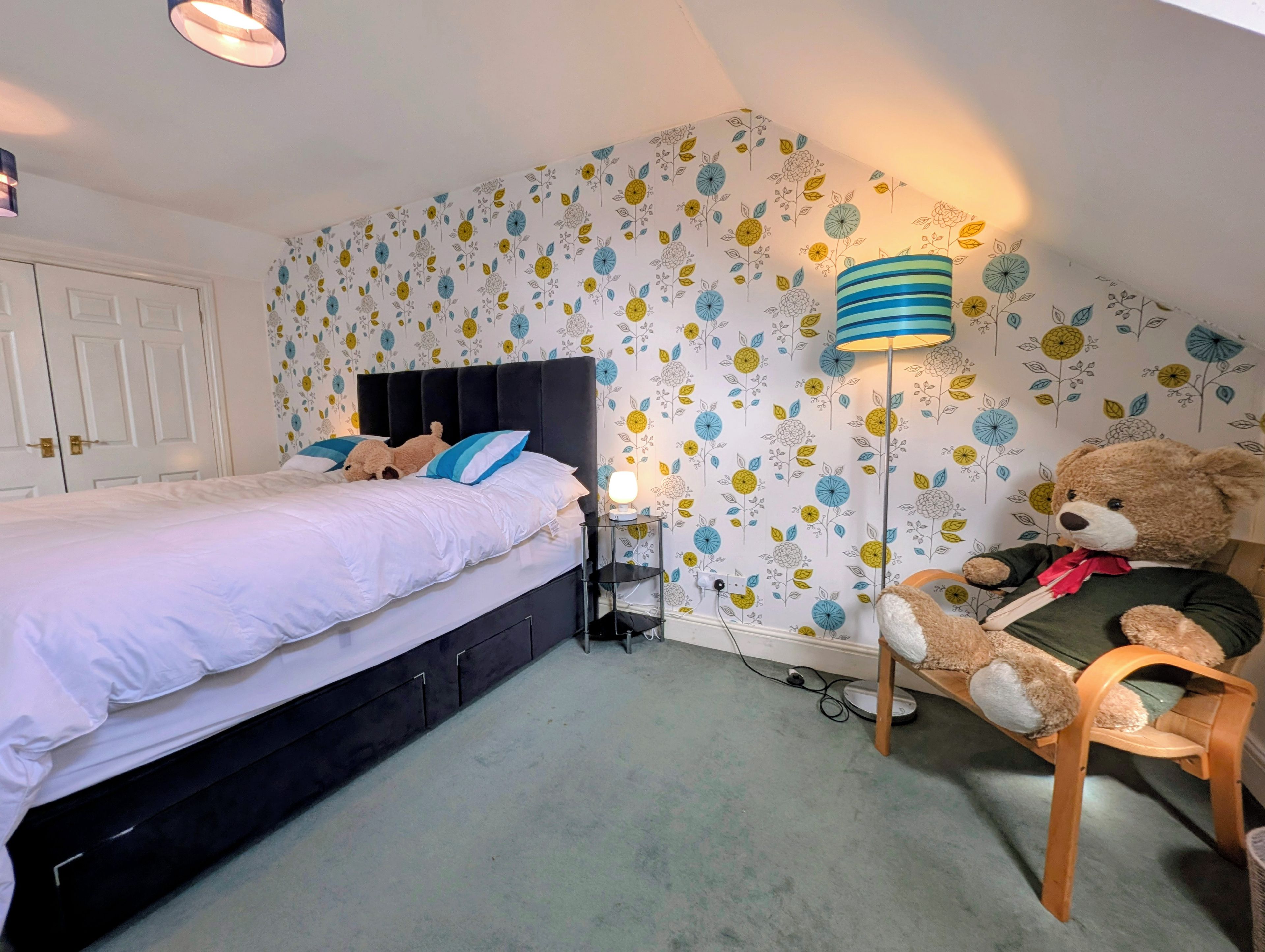 A bedroom with a bed, a nightstand, a lamp, a teddy bear, and a wall with floral wallpaper.