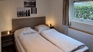 A double bed with white bedding, two bedside tables with lamps, a window with curtains, and wall art above the bed. - Holiday Home in Brunssum With Terrace (Brunssum)