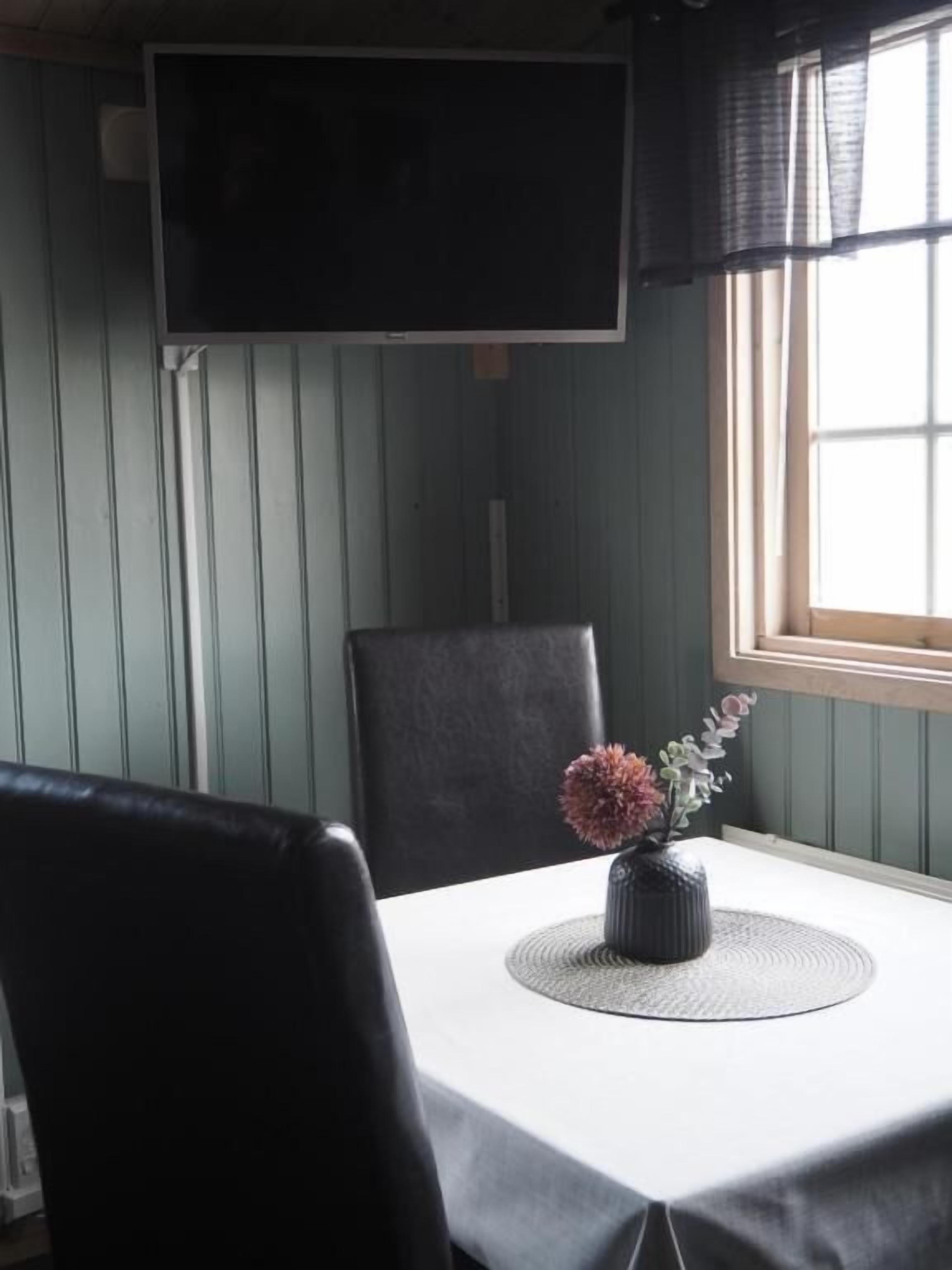 Ein Essbereich mit Tisch, Stühlen, einer Blumenvase, einem Fernseher und einem Fenster mit Vorhängen.