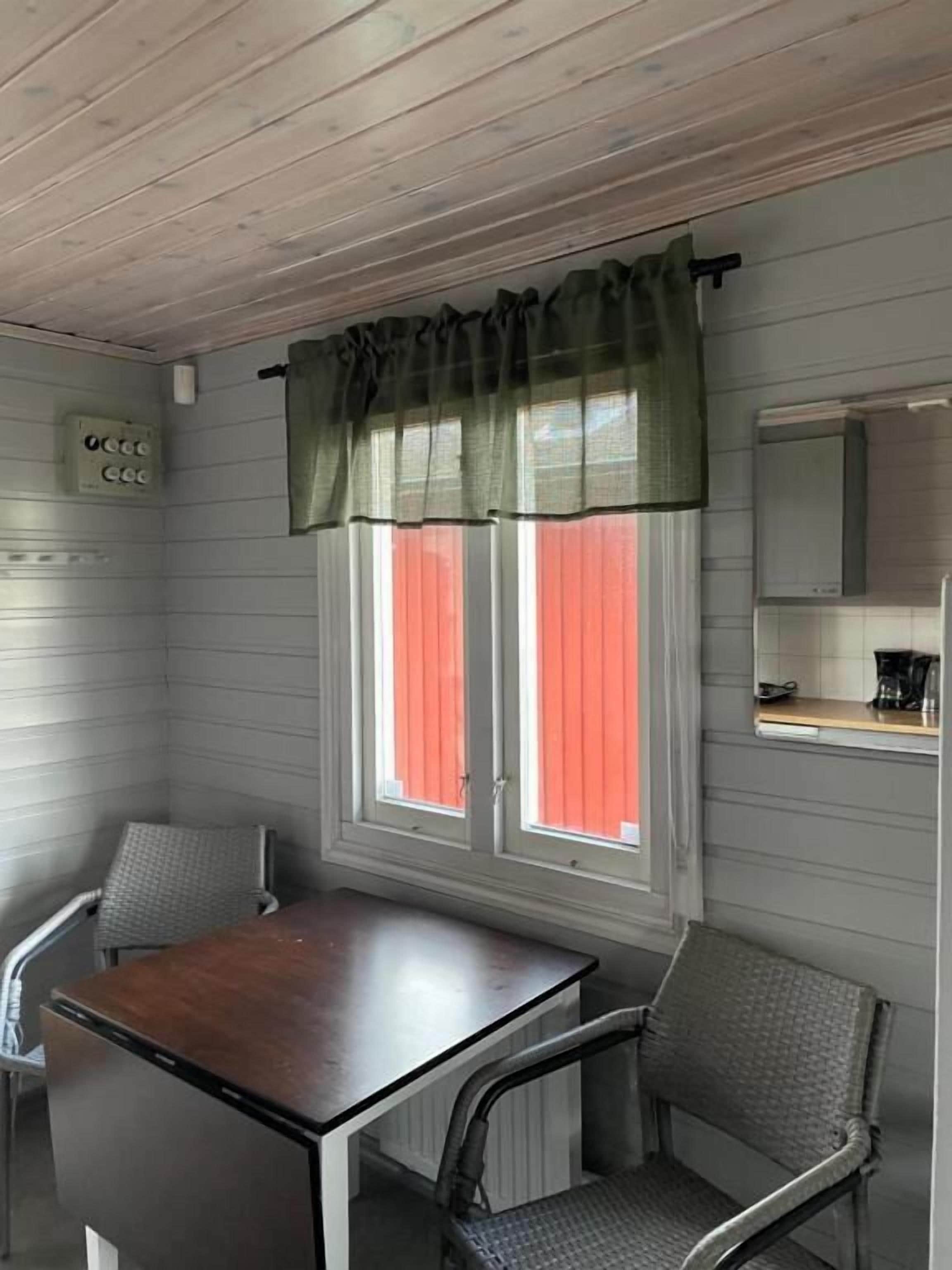 Una habitación con techo de madera, una ventana con cortinas verdes, una mesa, dos sillas y una pequeña zona de kitchenette.