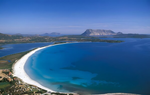 An aerial view of a coastal area with a beach, clear blue waters, and a prominent mountain in the background.