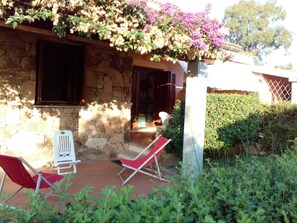 Ein Steinhaus mit einer überdachten Terrasse, zwei roten Stühlen und einem Garten mit grünen Sträuchern und violetten Blumen.