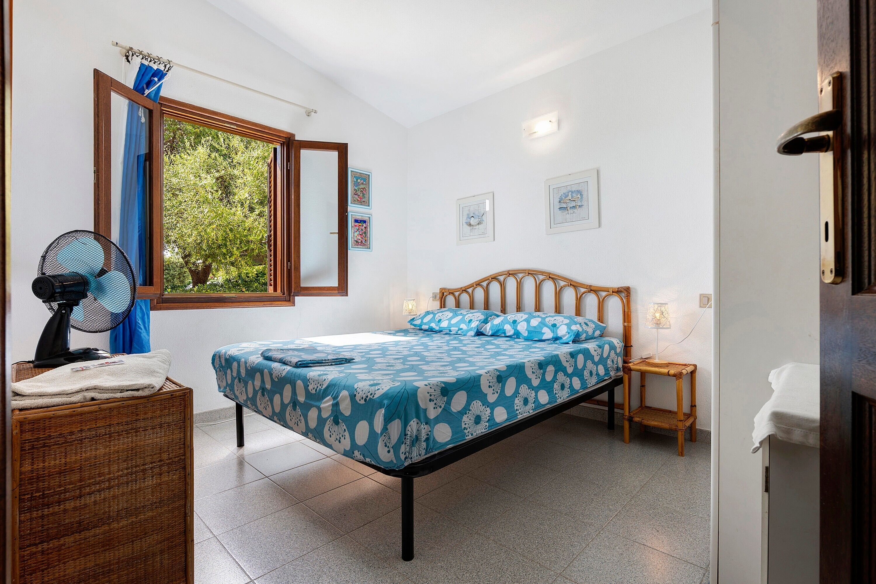 A bedroom with a bed, a standing fan, a window with curtains, and framed pictures on the wall.
