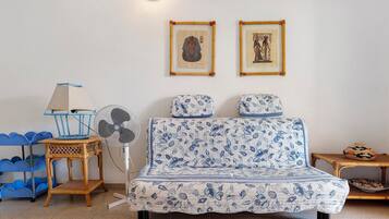 Een kamer met een blauwe gestreepte bank, een klein tafeltje met een ventilator, een rieten stoel en twee ingelijste foto's aan de muur.