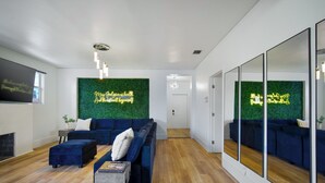 Ein modernes Wohnzimmer mit einer blauen Sofagarnitur, einem kleinen Couchtisch, einer grünen Wand mit einem Neonlicht und einem großen Spiegel.