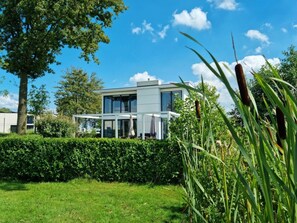 A modern two-story house with a glass facade, surrounded by a well-maintained hedge and tall grasses. - Cube Magnifique 10 (Nijkerk)