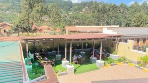 Un espacio de estar techado al aire libre con mesas y sillas, rodeado de vegetación y vistas a un paisaje montañoso.