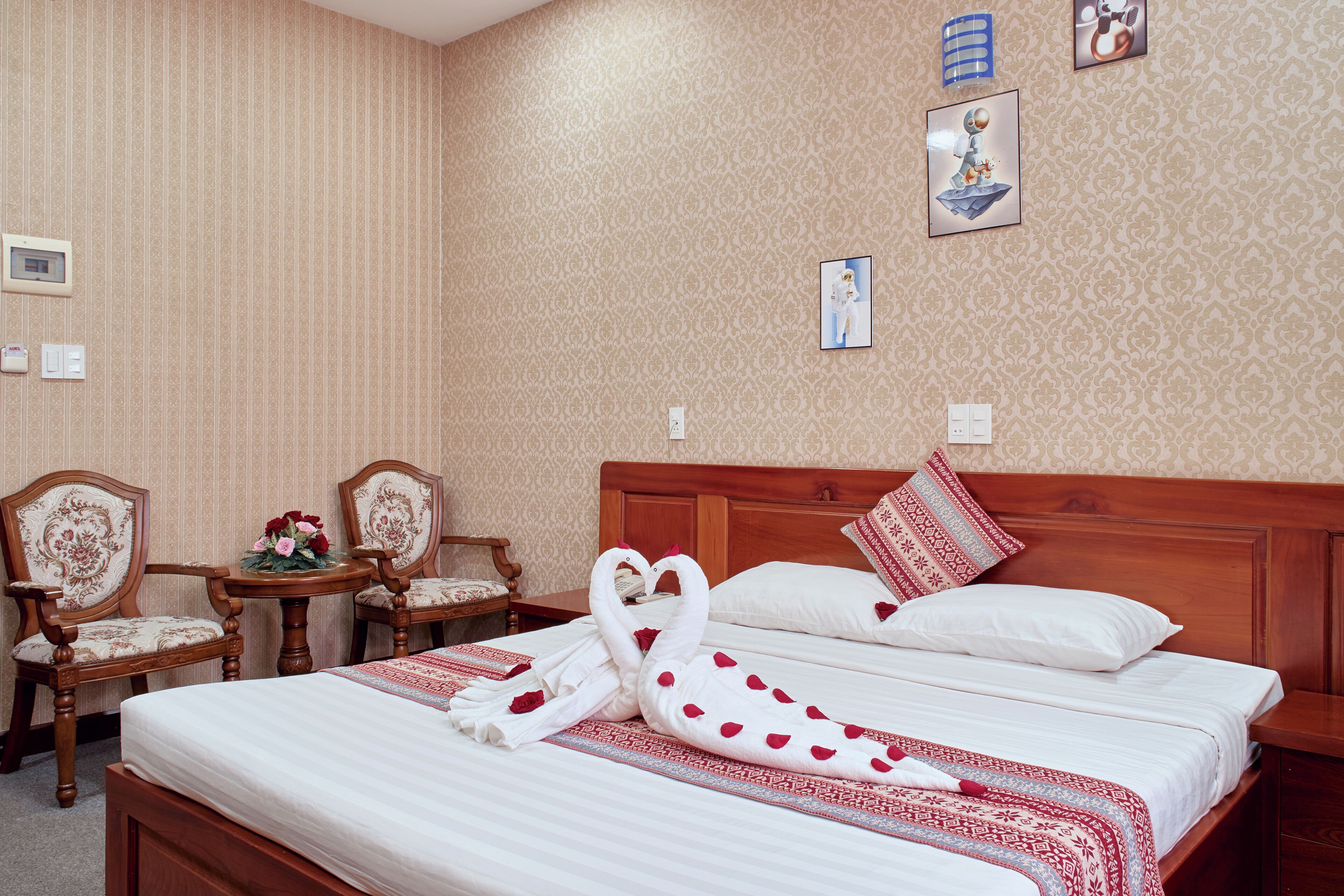 A hotel room with a neatly made bed, two chairs, a small table with flowers, and a wall clock.