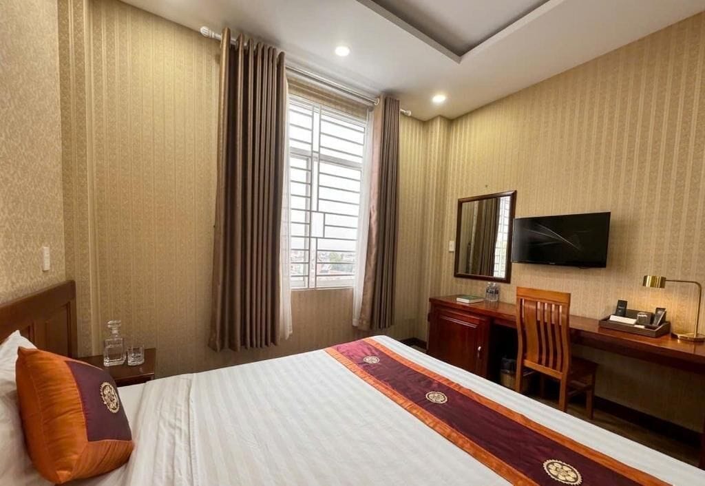 Una habitación de hotel con cama, escritorio, silla, televisor y ventana con cortinas.