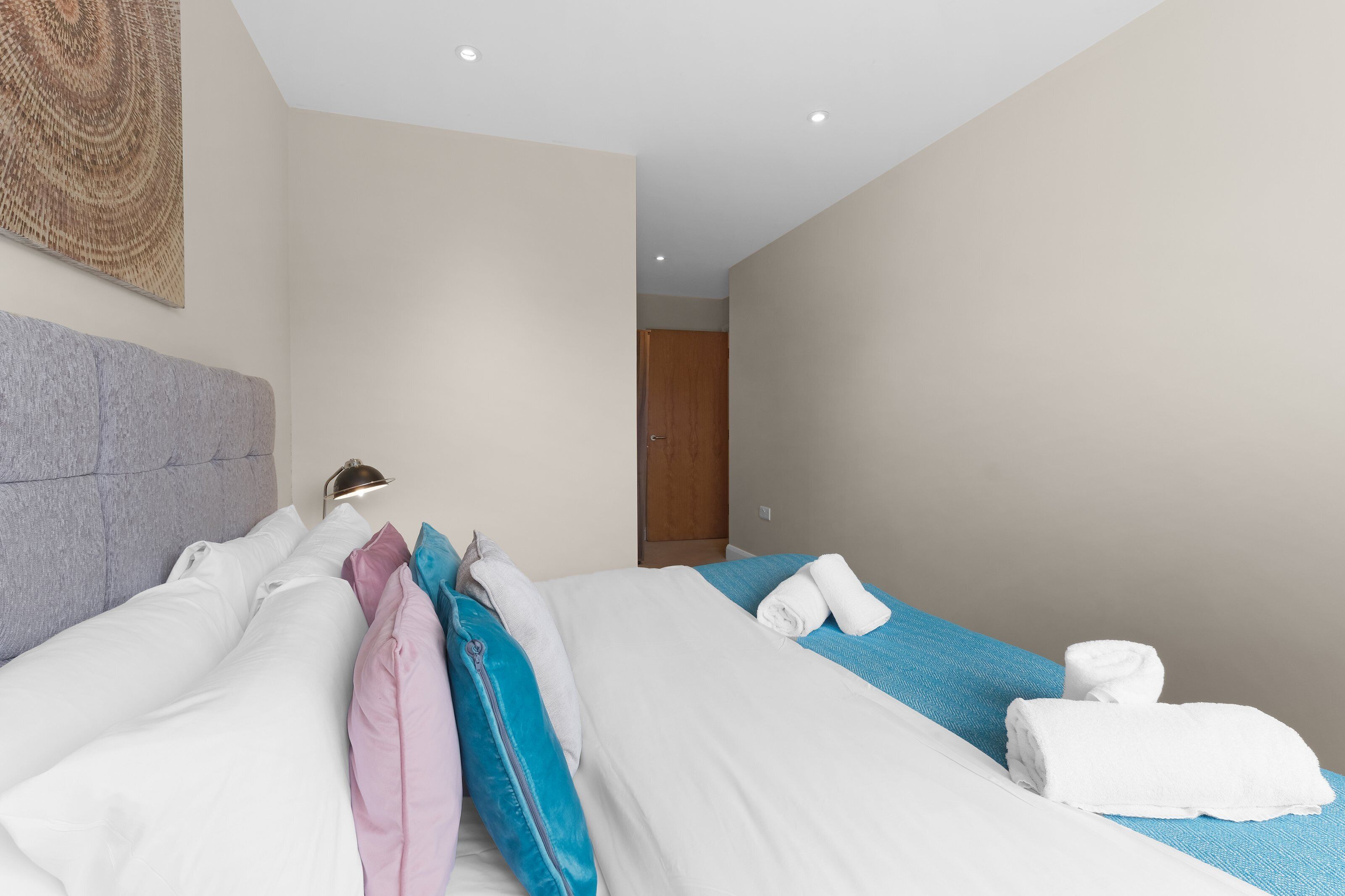 A hotel room with two beds, a headboard, a wall-mounted lamp, and a wooden door in the background.