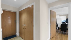 A hallway with wooden doors, a black chair, and a kitchen area in the background. - Heathrow Living 2BDR 2BA Apt 02-360stays (Stanwell)