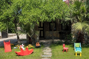 A garden with a stone pathway, colorful children's play equipment, and a stone building in the background. - Antik Manastır İda  (Edremit)