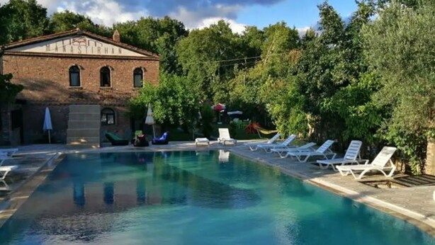 Una piscina con sillas de descanso y un edificio de ladrillo con el cartel 'MANASTIRHAN' al fondo.